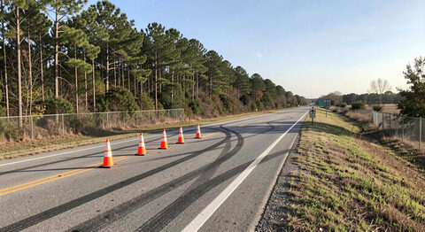 A two-lane Mississippi highway shoulder with skid marks and a set of traffic cones, pine trees along the fence line, clear sky.