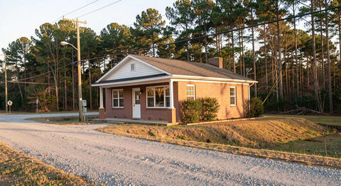 A rural Mississippi crossroads with a small brick office building, a gravel shoulder and pine woods behind, late-day sun.